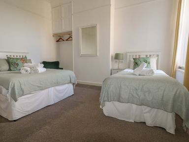 A bedroom with two beds each with green blankets and floral pillows a small open closet with hangers a chair a mirror and a bedside table with a lamp at Blue Yonder in Weymouth