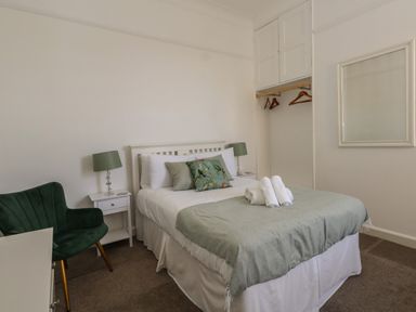 A bedroom with a bed covered with a blanket and pillows bedside tables with lamps a green chair an open closet with hangers and a wall mirror at Blue Yonder in Weymouth