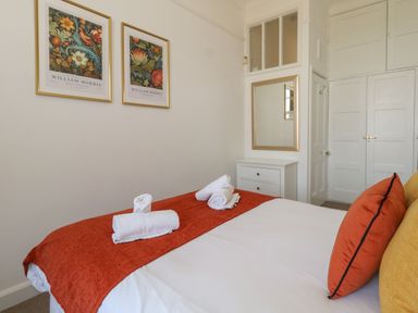 A bedroom with a bed covered with white sheets and an orange throw with rolled white towels on top two framed floral prints on the wall above a white chest of drawers and a mirror on the wall at Blue Yonder in Weymouth