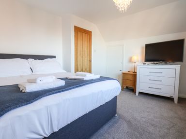 A bedroom with a bed with white linens and folded towels on it a wooden door a white dresser with a TV on top and a wooden nightstand with a lamp at Meadowfields in Whitby