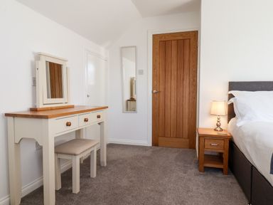 A bedroom with a dressing table, stool, bedside table with lamp, bed, wooden door, and wall mirror at Meadowfields in Whitby