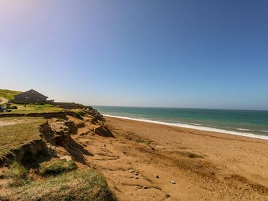 A sandy beach with cliff erosion next to a wooden building and the ocean at Alice's Cottage in Burton Bradstock
