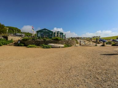 An outdoor area with a gravel surface a small building with green tents a stone wall a boat on the ground and a gate with cars and people beyond at Alice's Cottage in Burton Bradstock