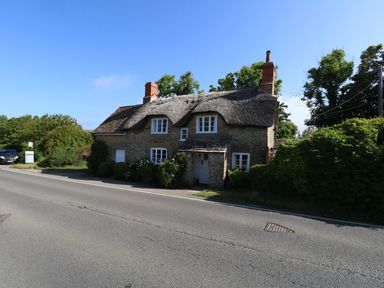 A stone house with a thatched roof and two chimneys next to a road with greenery around Alice's Cottage Burton Bradstock