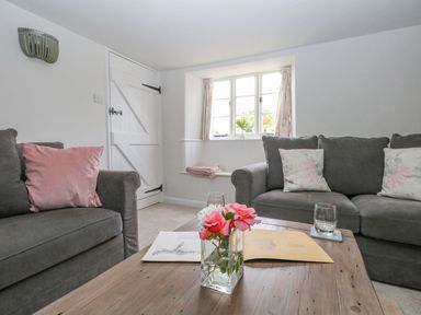 A living room with grey sofas pink and floral cushions a wooden coffee table with flowers and papers by a window with floral curtains at Alices Cottage in Burton Bradstock