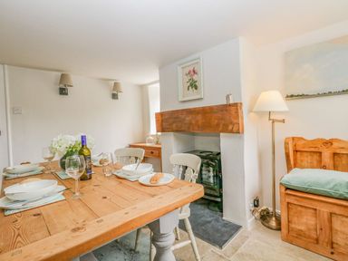 A dining area with a wooden table set with plates and glasses near a small fireplace and a wooden bench at Alices Cottage in Burton Bradstock
