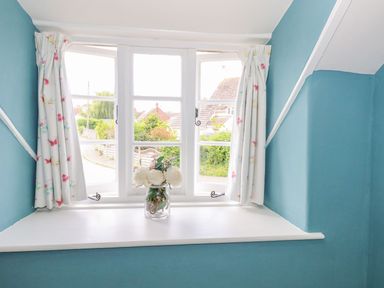 A window with white frame and floral curtains a glass jar with white flowers on the windowsill at Alices Cottage in Burton Bradstock