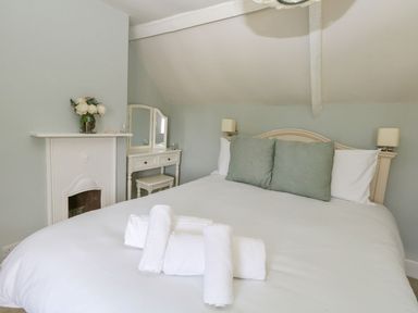 A bedroom with a bed with white linens and green pillows a white vanity with mirror and a small fireplace with flowers on top at Alices Cottage in Burton Bradstock