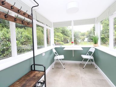A sunroom with large windows, two white folding chairs, a small wall-mounted table, a plant, and a wooden coat rack Alice's Cottage Burton Bradstock