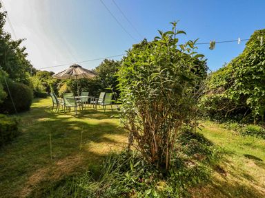 A garden with a dining table and chairs under a parasol surrounded by bushes at Alices Cottage in Burton Bradstock