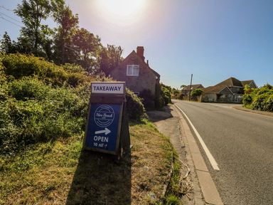 A roadside sign advertising takeaway for Hive Beach Cafe near houses and greenery in Burton Bradstock