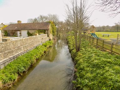 A stream with grass and trees alongside a stone wall and a playground with slides at Alices Cottage in Burton Bradstock