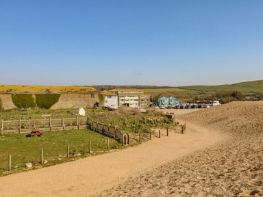 A sandy path with a fenced grassy area and buildings in the background at Alices Cottage in Burton Bradstock