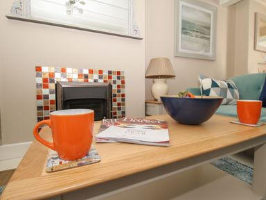 A living room with a wooden coffee table holding orange mugs magazines and a bowl in front of a fireplace at Little Gull Cottage in Weymouth