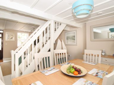 A dining area with a wooden table and white chairs next to a white staircase and a sideboard with a mirror at Little Gull Cottage in Weymouth