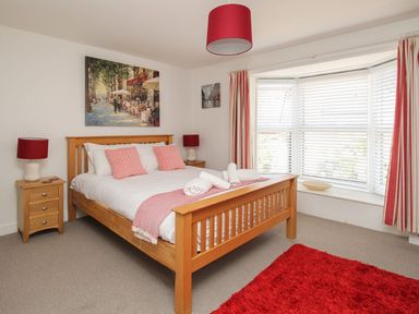 A bedroom with a wooden bed frame bedside tables with lamps and a bay window with blinds and red and white curtains at Little Gull Cottage in Weymouth