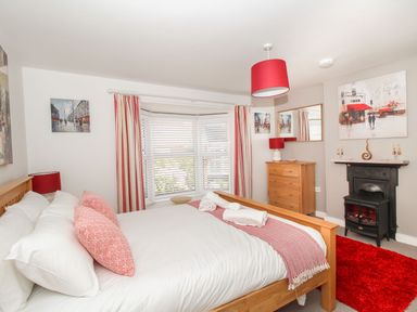 A bedroom with a wooden bed with white bedding and red pillows a wooden dresser a black fireplace a red rug and a window with striped curtains at Little Gull Cottage in Weymouth