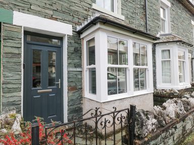 The exterior of a stone house with a dark door and white framed bay windows behind a black metal gate with red berries and rock wall at Forty Five in Keswick