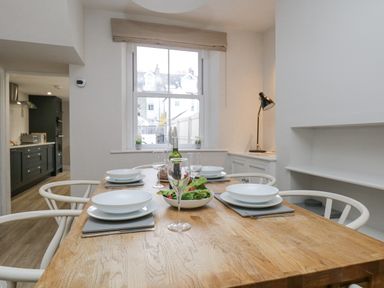 A dining table set with white plates bowls wine glasses and a salad bowl in a room with a window and kitchen visible at Forty Five in Keswick