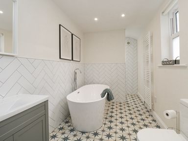 A bathroom with a freestanding bathtub, patterned floor tiles, white herringbone wall tiles, a grey vanity unit, and framed sketches on the wall at Forty Five in Keswick