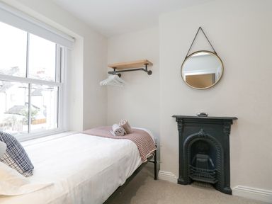 A small bedroom with a single bed near a window a round mirror above a black decorative fireplace and a wall shelf with hangers at Forty Five in Keswick