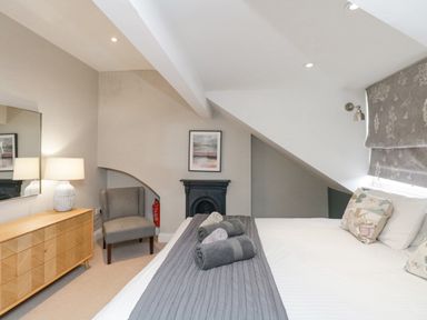 A bedroom with a bed with rolled towels a grey armchair a wooden dresser with a lamp and a small fireplace at Forty Five in Keswick