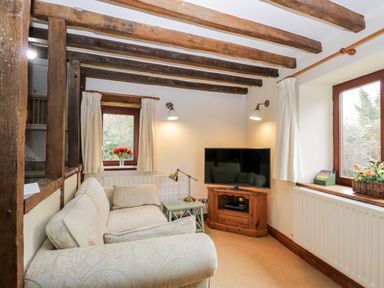 A small living room with a patterned sofa wooden beams on ceiling a TV on wooden stand and flowers on window sills at Well Cottage in Moreton-In-Marsh