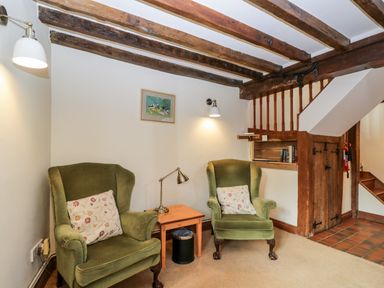 A sitting area with two green armchairs a small table and a lamp under wooden ceiling beams at Well Cottage in Moreton-In-Marsh