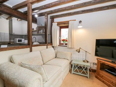 A living room with a patterned sofa a small side table with a lamp a window with curtains and flowers and a tv on a wooden stand at Well Cottage in Moreton-In-Marsh