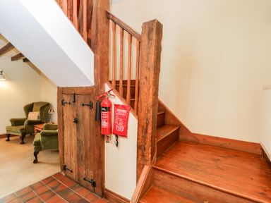 A wooden staircase next to a small wooden door and a fire extinguisher with a fire blanket in a hallway with a green armchair visible in the adjacent room at Well Cottage in Moreton-In-Marsh