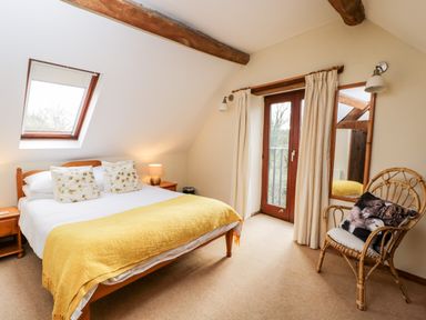 A bedroom with a bed covered with yellow blanket and floral pillows beside a window and door and a wicker chair with animal print cushion at Well Cottage in Moreton-In-Marsh