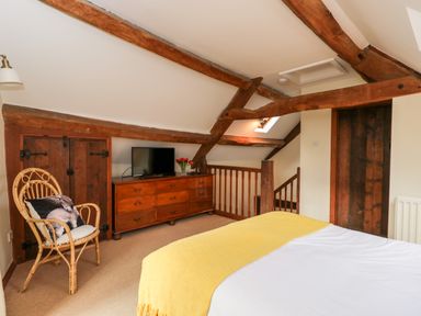 A bedroom with a bed covered in white and yellow bedding a wooden dresser a TV a wicker chair with a cushion and exposed wooden beams at Well Cottage in Moreton-In-Marsh