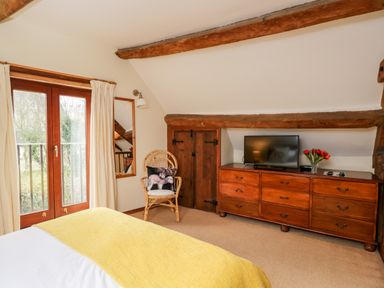 A bedroom with wooden beams a dresser with a television and flowers and a chair with a cushion at Well Cottage in Moreton-In-Marsh