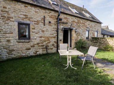 A stone cottage exterior with a small table and two chairs on a grassy lawn at Well Cottage in Moreton-In-Marsh