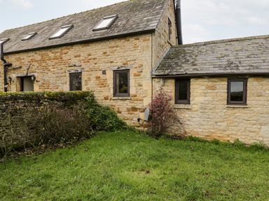 An exterior view of a stone house with multiple windows a stone wall and grass at Stable Cottage in Moreton-In-Marsh