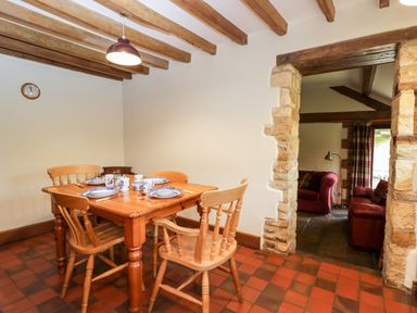 A dining room with a wooden table and chairs set with dishes next to a stone doorway leading to a sitting room at Stable Cottage in Moreton-In-Marsh