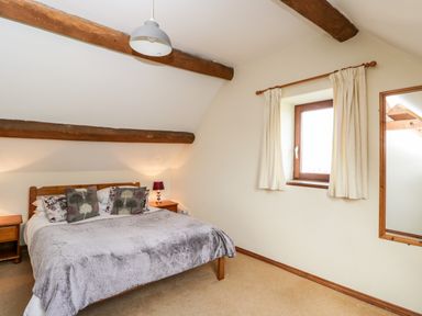 A bedroom with a wooden bed with pillows and a gray blanket two wooden bedside tables a window with curtains and a mirror on the wall at Stable Cottage in Moreton-In-Marsh