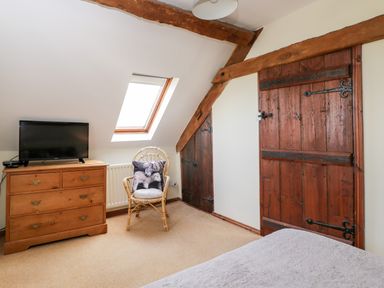 A bedroom with wooden doors a dresser with a television and a wicker chair with a cushion at Stable Cottage in Moreton-In-Marsh
