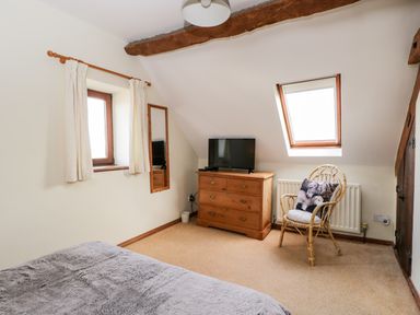 A bedroom with a bed a wooden dresser with a television a wicker chair with a cushion and two windows at Stable Cottage in Moreton-In-Marsh