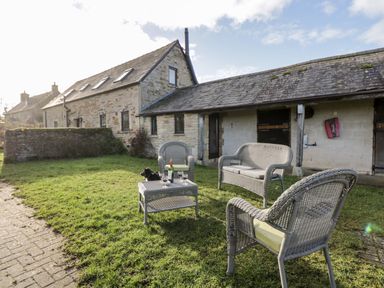 An outdoor seating area with wicker furniture and a dog on grass next to a stone building at Stable Cottage in Moreton-In-Marsh