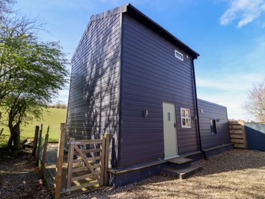 An exterior view of a dark grey metal sided building with a small beige door and steps surrounded by a wooden gate and gravel at The Stables in Tafarn-Y-Gelyn