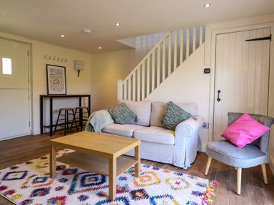 A living room with a beige sofa with green pillows a gray chair with a pink pillow a wooden coffee table and a colorful patterned rug at The Stables in Tafarn-Y-Gelyn