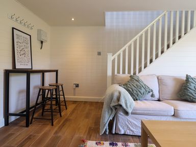 A living room with a sofa, two stools, a table, a framed picture, and stairs in the background at The Stables in Tafarn-Y-Gelyn