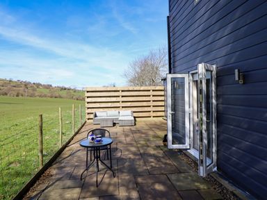 An outdoor patio with a small table and chair set and a cushioned sofa against a wooden fence at The Stables in Tafarn-Y-Gelyn
