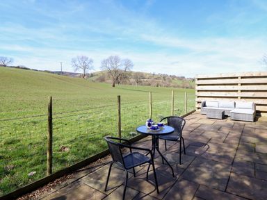 An outdoor patio with a table and two chairs and a seating area overlooking a grassy field at The Stables in Tafarn-Y-Gelyn