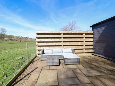 An outdoor patio with a gray wicker sectional sofa and a small table on tiled flooring next to a wooden fence and green field at The Stables in Tafarn-Y-Gelyn