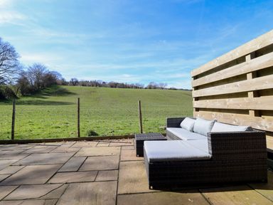 A patio with a wicker sofa and cushions overlooking a grassy field at The Stables in Tafarn-Y-Gelyn