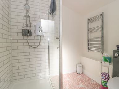 A bathroom with a shower area with white subway tile walls and a handheld showerhead a chrome towel radiator a small white trash bin and patterned pink floor tiles at The Potting Shed in Combe near Langport