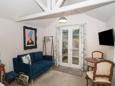 A room with a blue sofa a small round table with two chairs a wall-mounted TV and glass doors with curtains at The Potting Shed in Combe near Langport