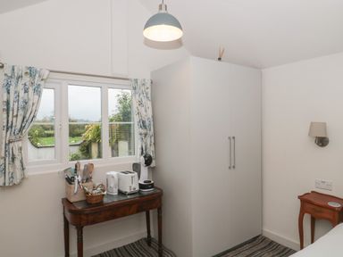 A room with a window with floral curtains a wooden table with kitchen appliances a white wardrobe and a small wooden side table at The Potting Shed in Combe near Langport